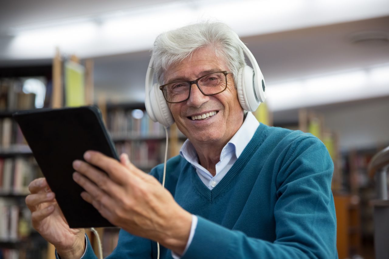 The Art of Drawing Readers In: Your attractive post title goes here Senior man wearing headphones using a tablet in a library. Happy expression showcases enjoyment of technology.